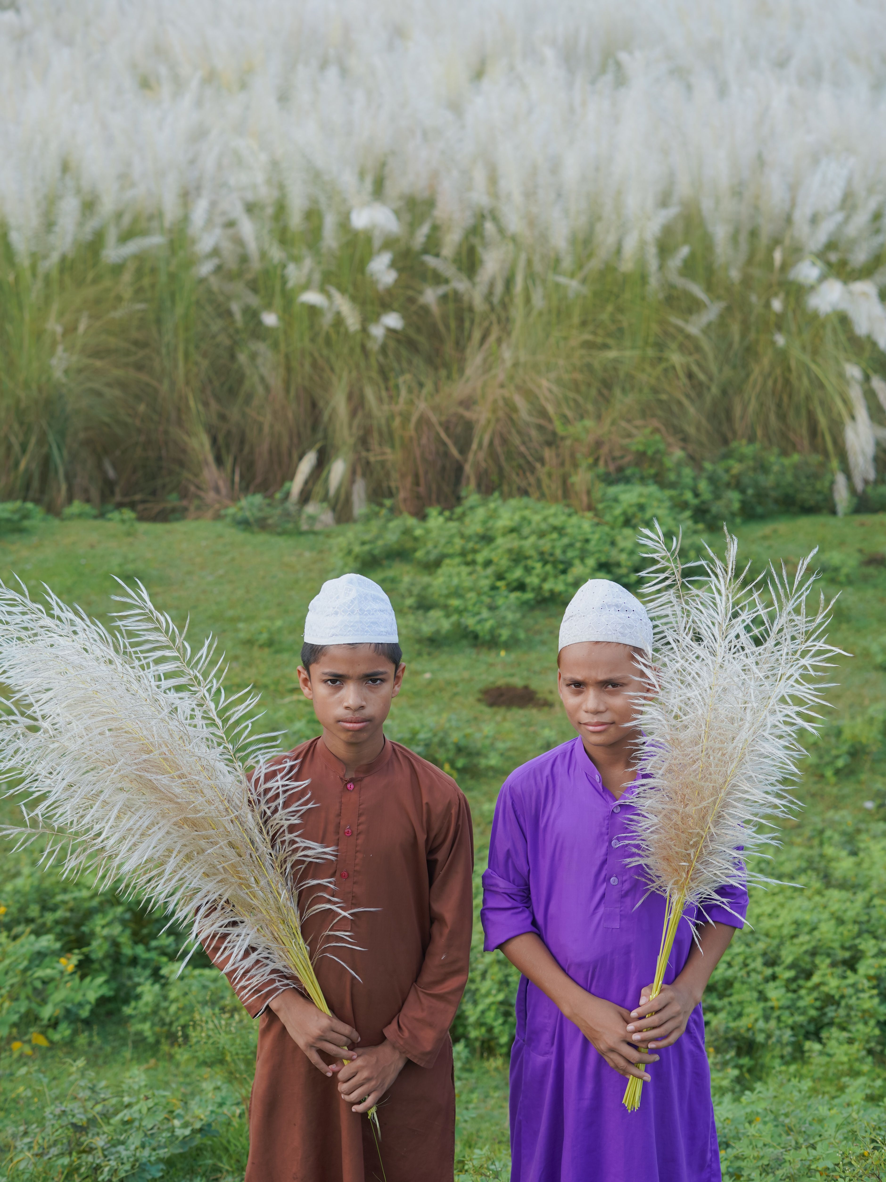 Two boys in traditional attire holding pampas grass in a lush Bangladeshi field.