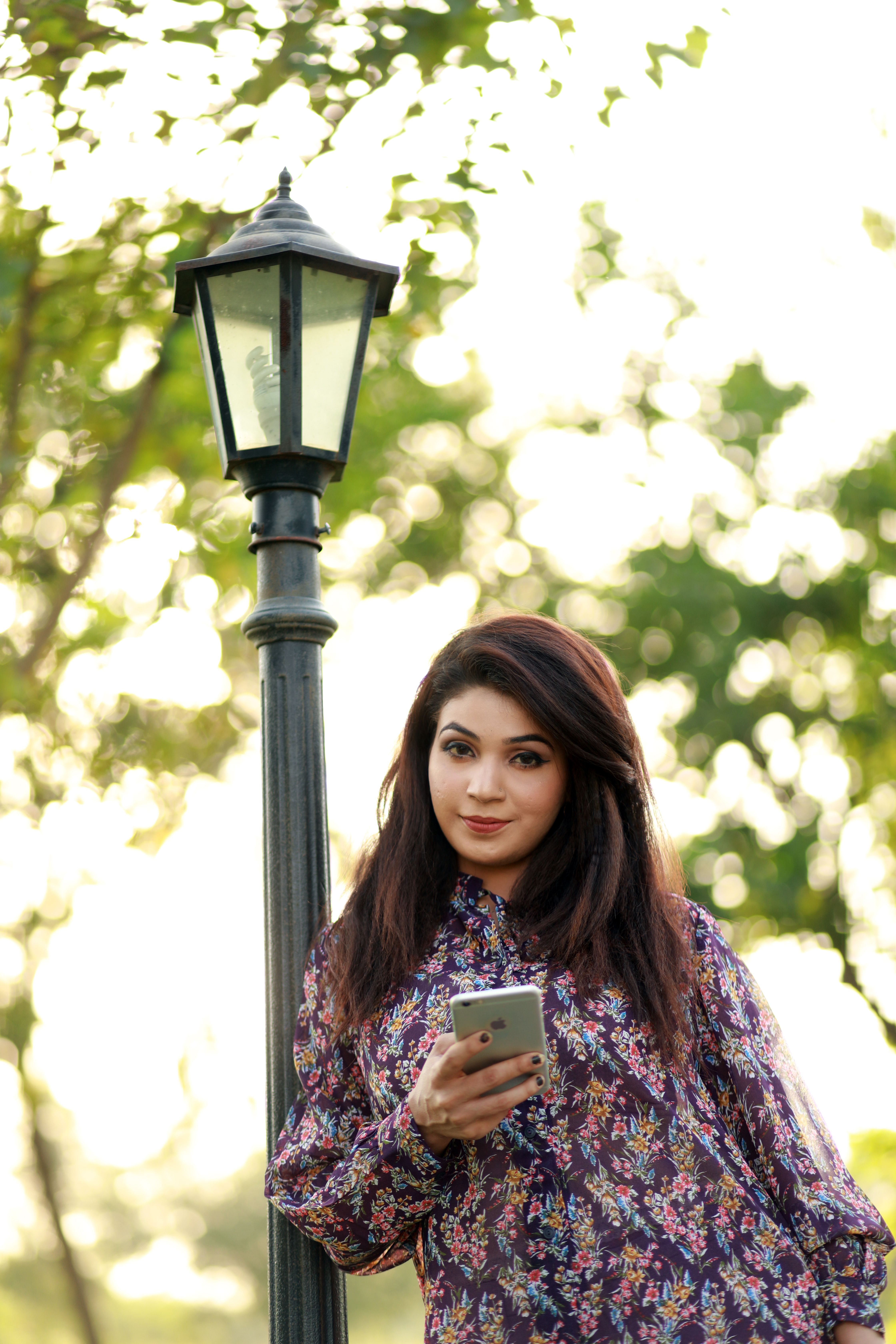 Asian woman in floral attire using a smartphone by a street lamp outdoors.
