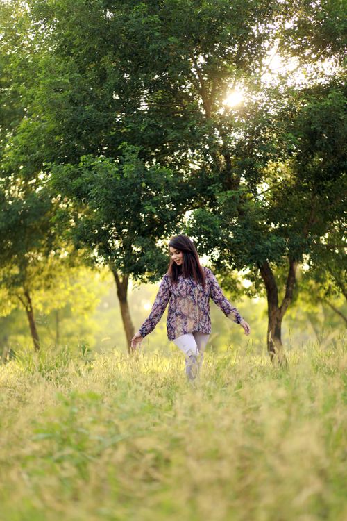 Free A woman walking through a sunlit grassy field surrounded by lush green trees, embodying serenity. Stock Photo
