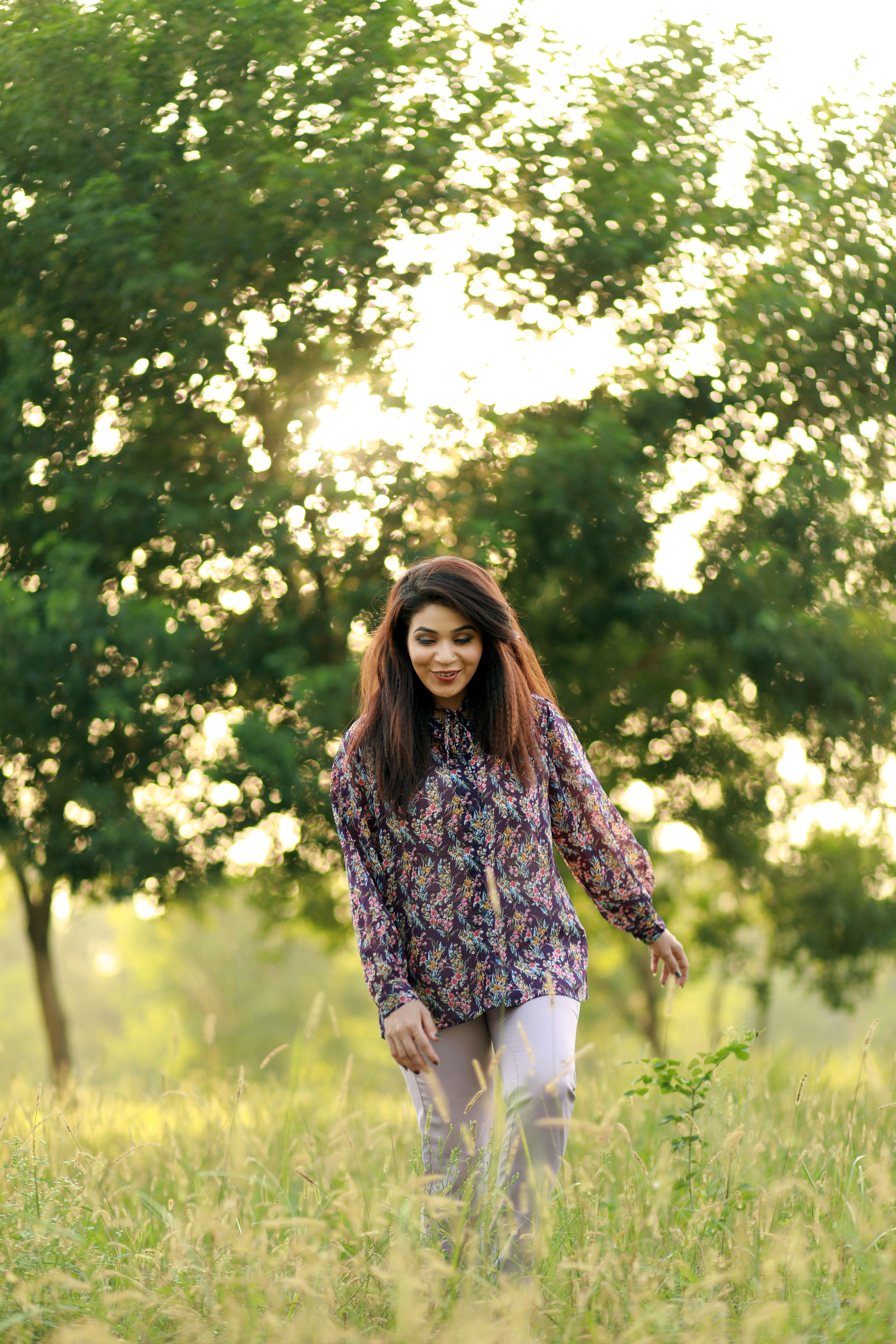A woman in floral attire walks through a sunlit grassy field, surrounded by trees.