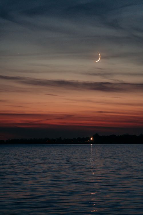 Free A tranquil evening scene of a crescent moon over calm ocean waters with a vibrant sunset sky. Stock Photo