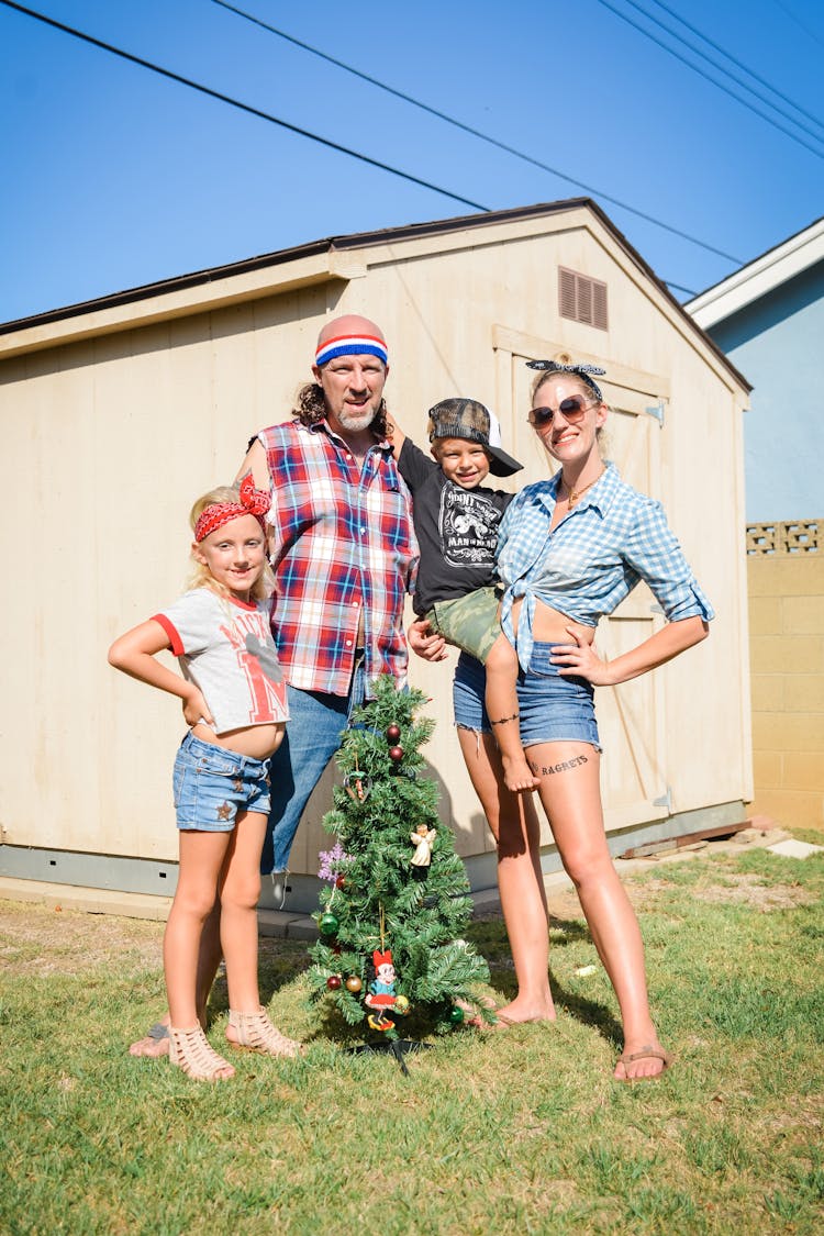 Parents With Two Children Standing Next To A Tiny Christmas Tree On The Backyard In Summer