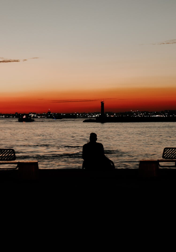 Silhouette Of Person Sitting On Bench Near Body Of Water During Sunset