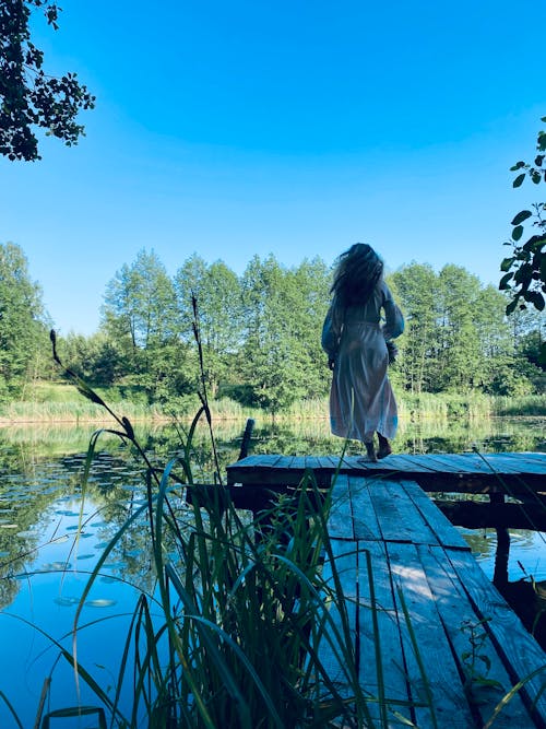A Woman in White Dress Standing on a Wooden Dock Near Body of Water and Trees