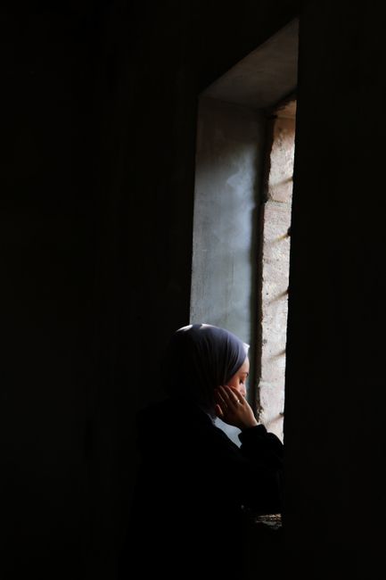 Silhouette of a woman in headscarf gazing out of a dimly lit window.