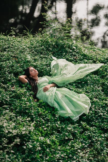 A woman in a green dress lies peacefully on a foliage-covered ground, surrounded by nature.