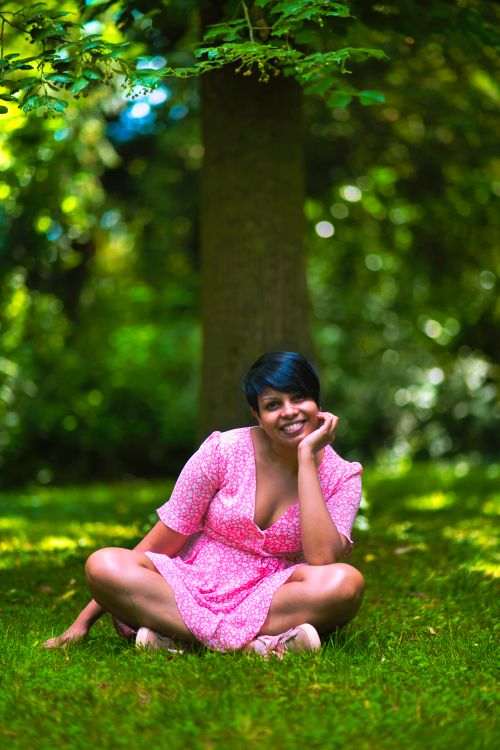 Free A smiling woman in a pink dress poses happily outdoors amidst lush greenery. Stock Photo