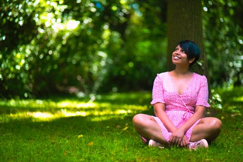 Free Asian woman in pink dress sitting happily on grass under trees in daylight, enjoying nature. Stock Photo