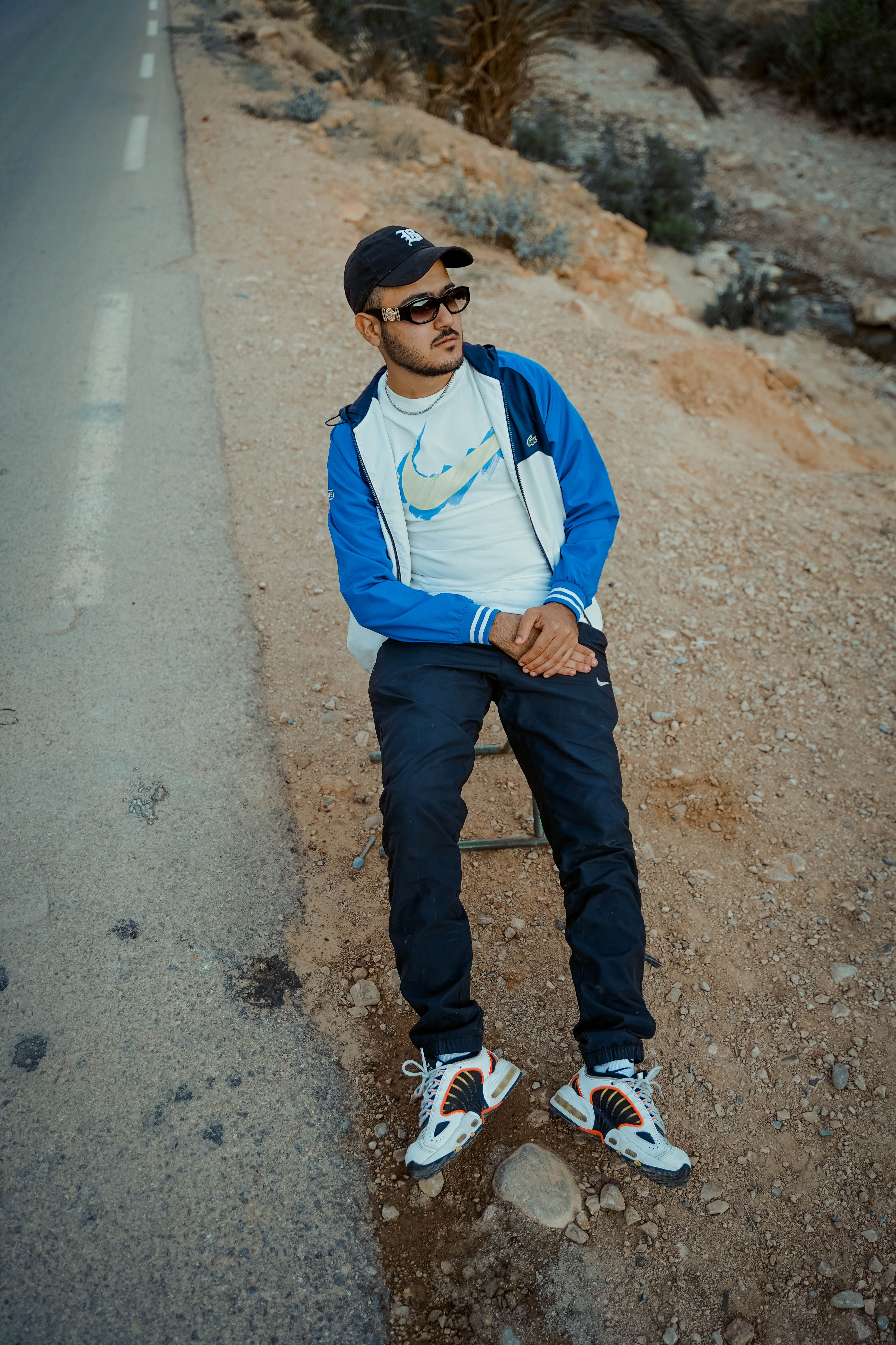 A stylish man in casual streetwear sits beside a desert road in Biskra, Algeria.