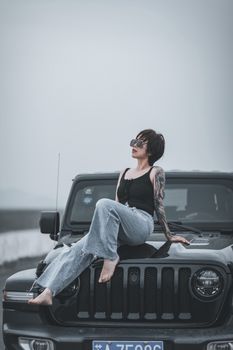 Stylish woman with tattoos and sunglasses sitting on an SUV hood outdoors, showcasing fashion and confidence.