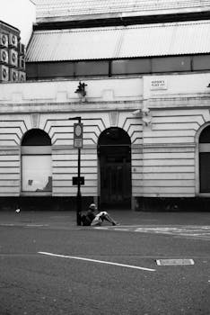 A black and white street photo of a person sitting by a building in London, England.