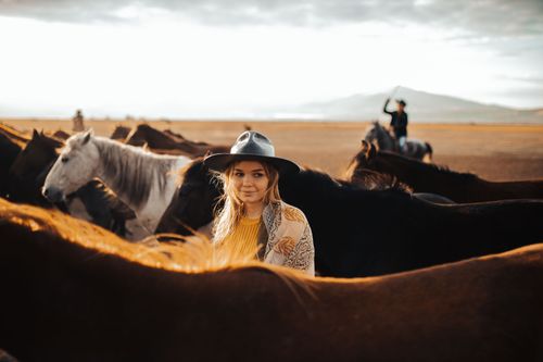 Free A woman with blonde hair and fedora hat surrounded by horses in a vast landscape with a cowboy in the background. Stock Photo
