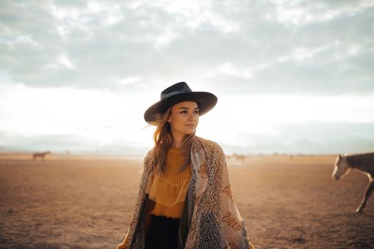 A woman in a cowboy hat stands in a sunny desert with horses in the background.