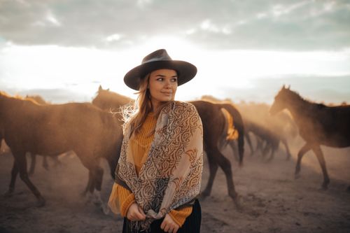 Free Woman in a fedora stands amidst a herd of horses at sunset, exuding a serene and adventurous vibe. Stock Photo