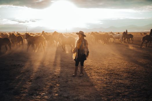 A woman standing amidst a herd of horses at sunset, enveloped in dust, in a vast open field.