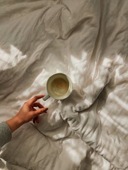 A serene image of a hand holding an empty mug on a bed, bathed in soft morning light and shadows.