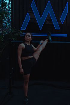 Woman stretches leg on gym bar under dark lighting. Athletic and focused, wearing sportswear.
