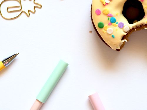 Free Flat lay of a donut with icing and sprinkles, surrounded by pastel pens and a paperclip on a white background. Stock Photo