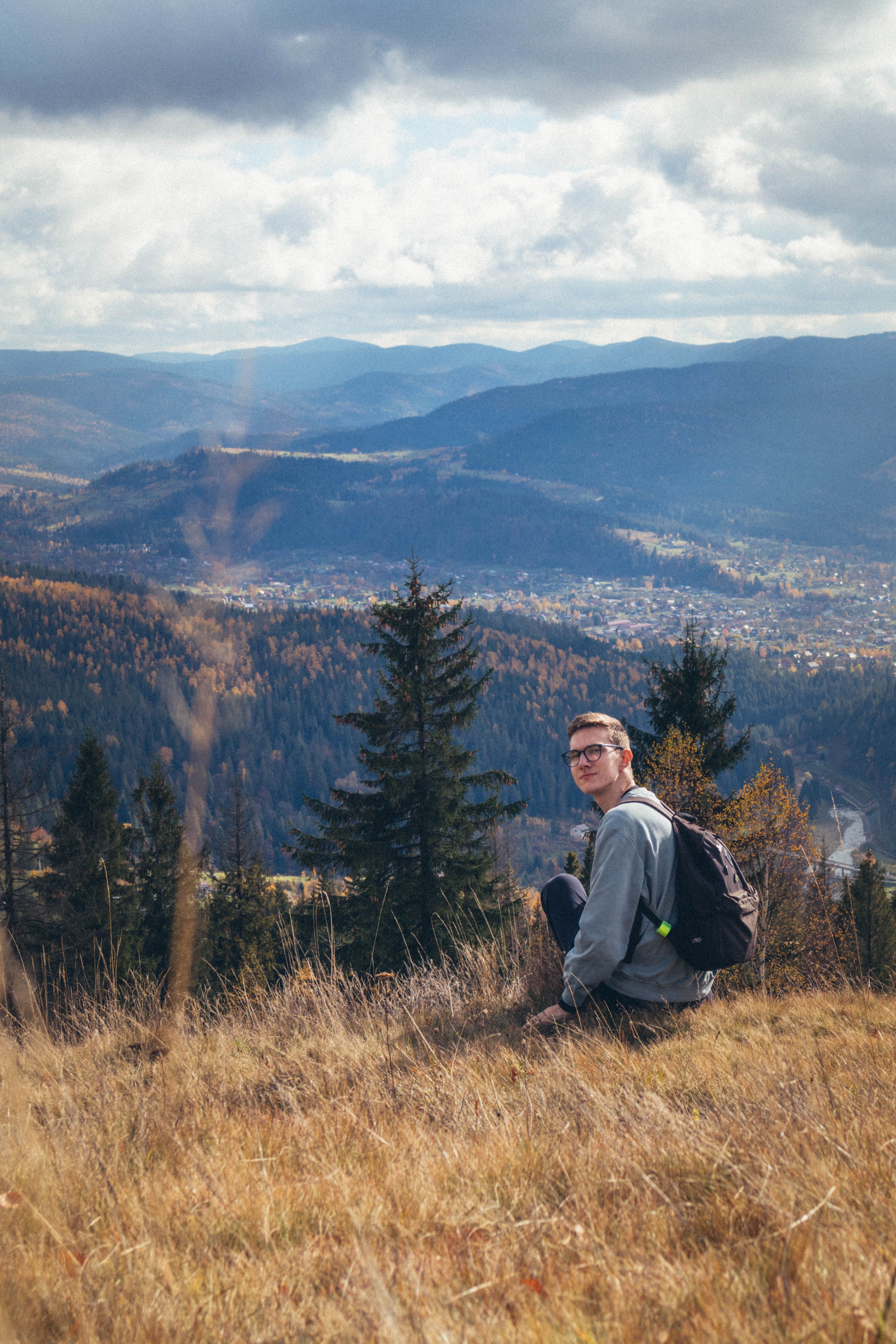 Man with backpack sitting on a hill, overlooking an autumn mountain landscape in daylight.