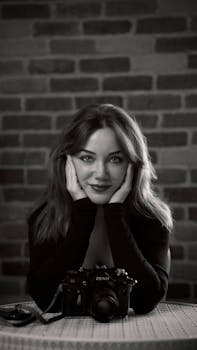 Black and white portrait of a woman sitting at a table, hands on face, beside a camera.