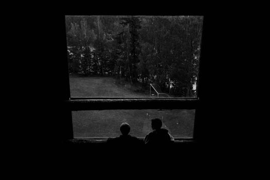 Two individuals silhouetted against a forest view from a window, in black and white.