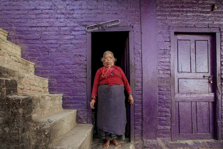 Old Woman Standing In House Doorway 