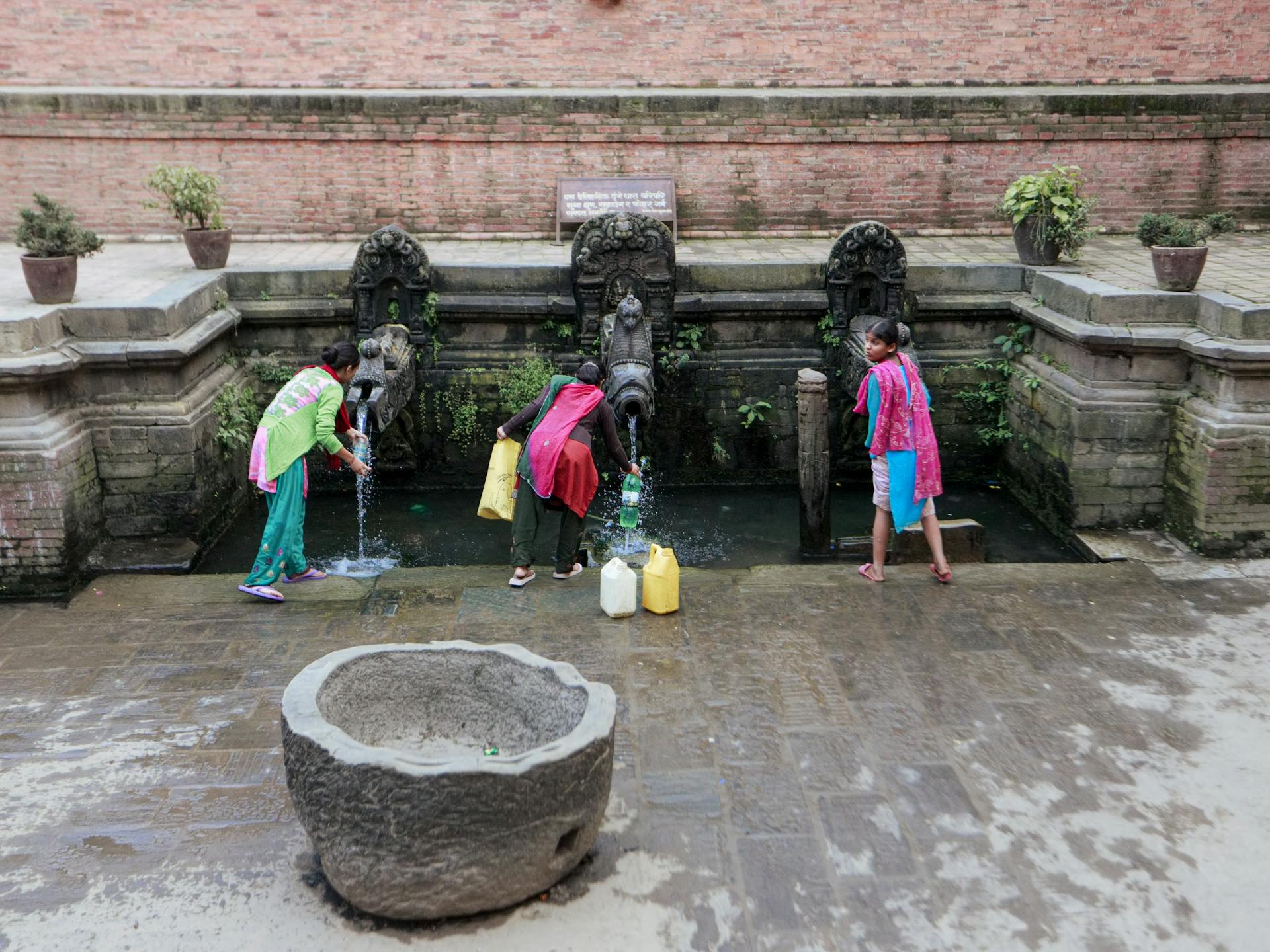 Three women filling containers at an ancient decorative water fountain in an outdoor urban setting.