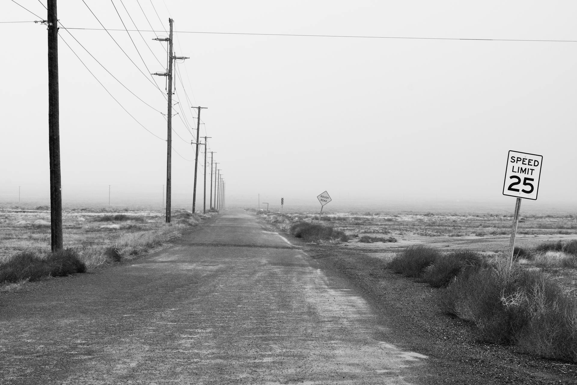 A deserted road with power lines and a speed limit sign in a flat desert landscape.