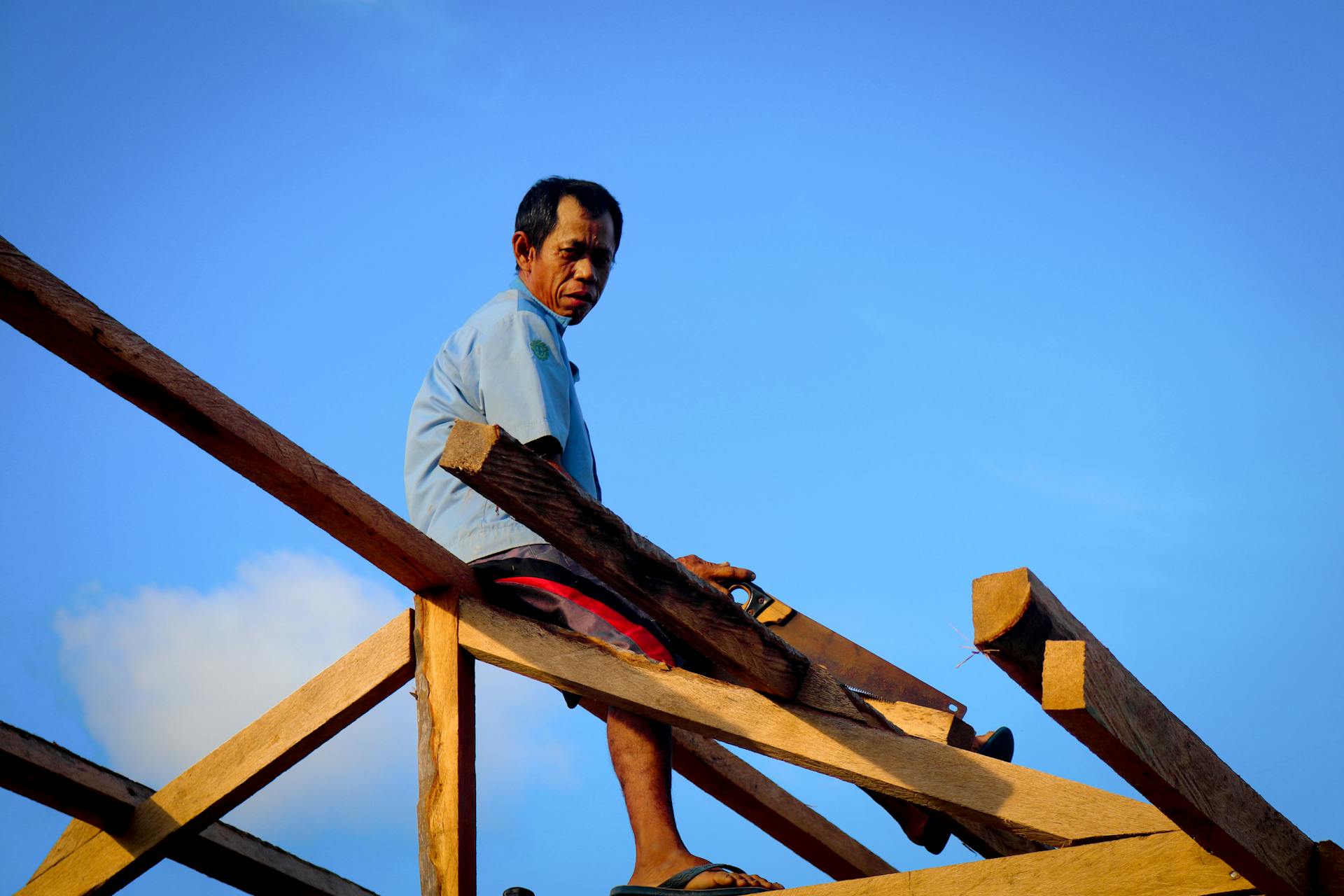 Asian carpenter skillfully sawing wooden beams on a roof under a bright blue sky.