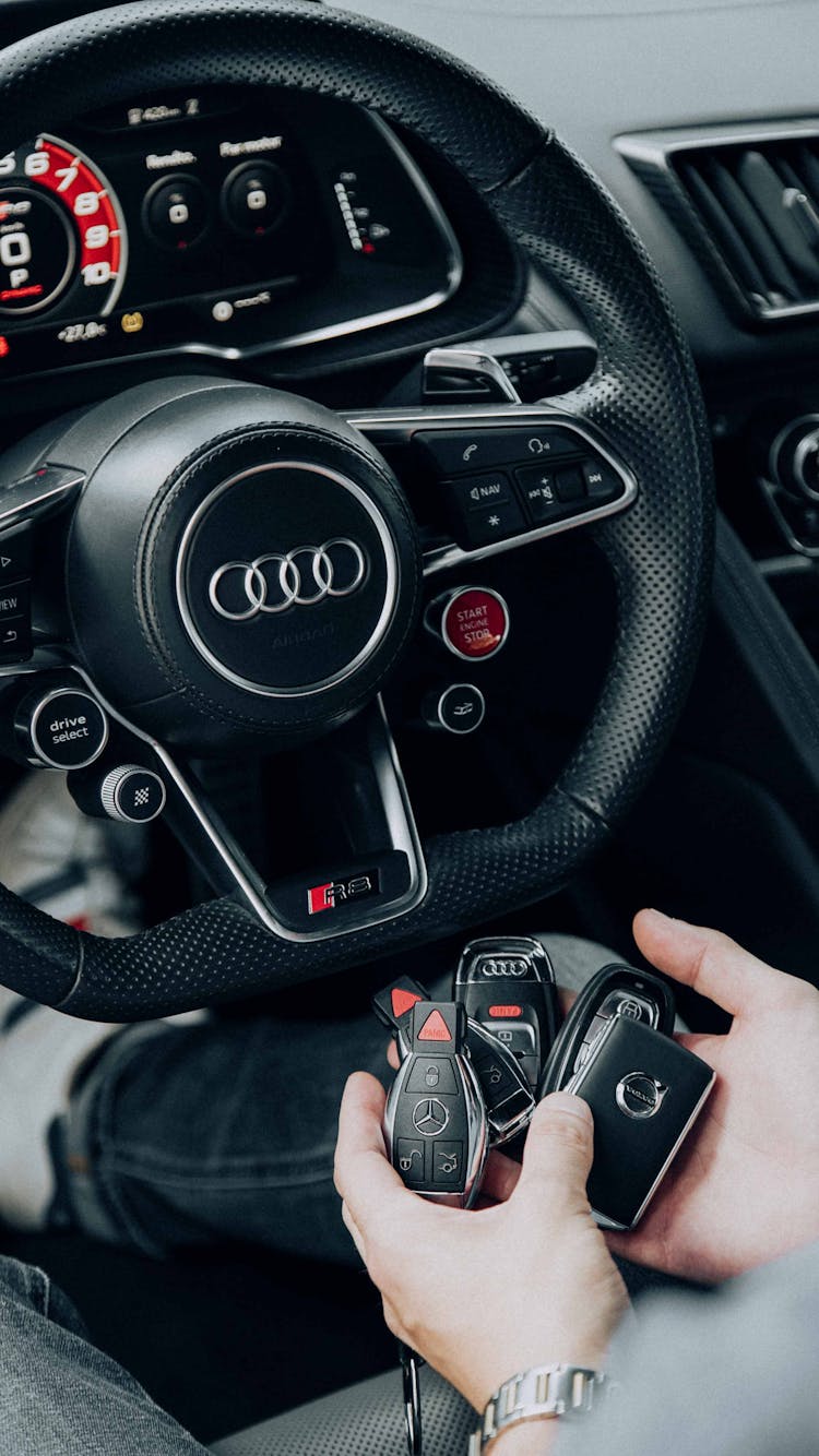 Person Holding Car Keys Sitting Behind A Black Leather Steering Wheel