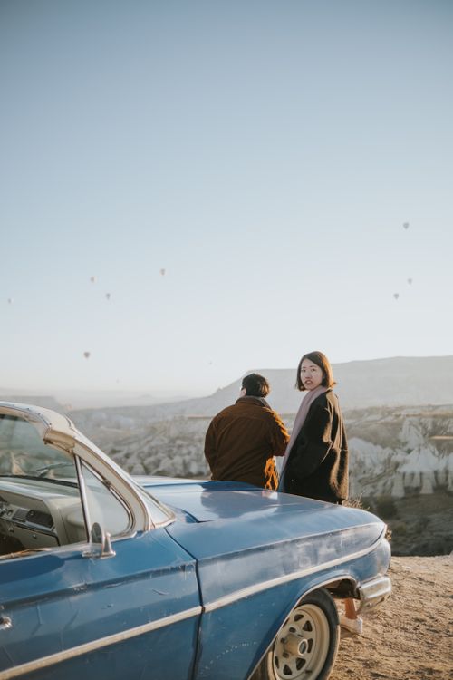 Free A couple stands next to a vintage car, enjoying a scenic view of hot air balloons in Cappadocia, Turkey. Stock Photo