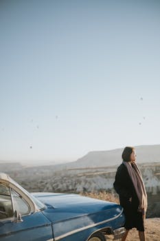 Woman in scarf and coat stands by vintage car in tranquil landscape with hot air balloons.