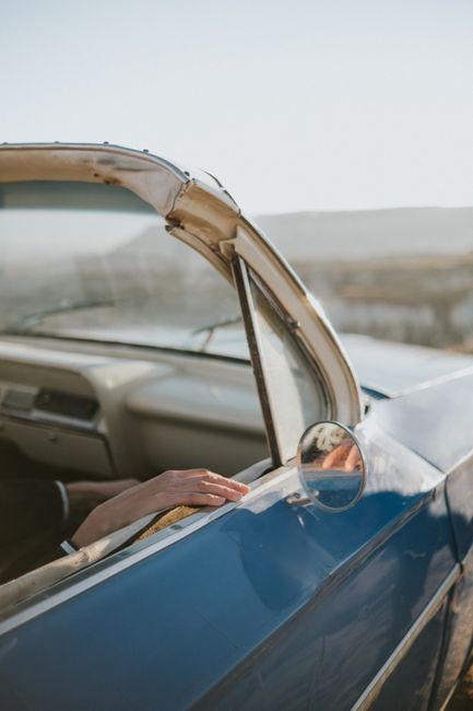 Close-up of a hand resting on a vintage blue convertible's window with an outdoor scenic backdrop.