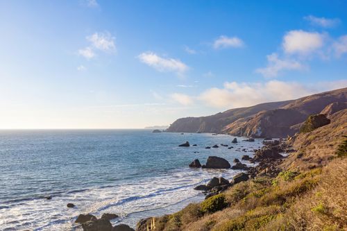 Free Beautiful view of a rocky coastline with waves and hills under a clear blue sky. Stock Photo