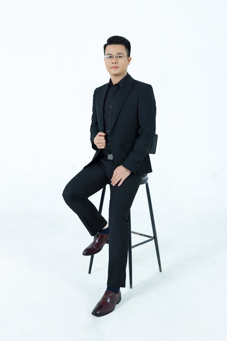 A Man In A Black Suit Sitting On A Chair 