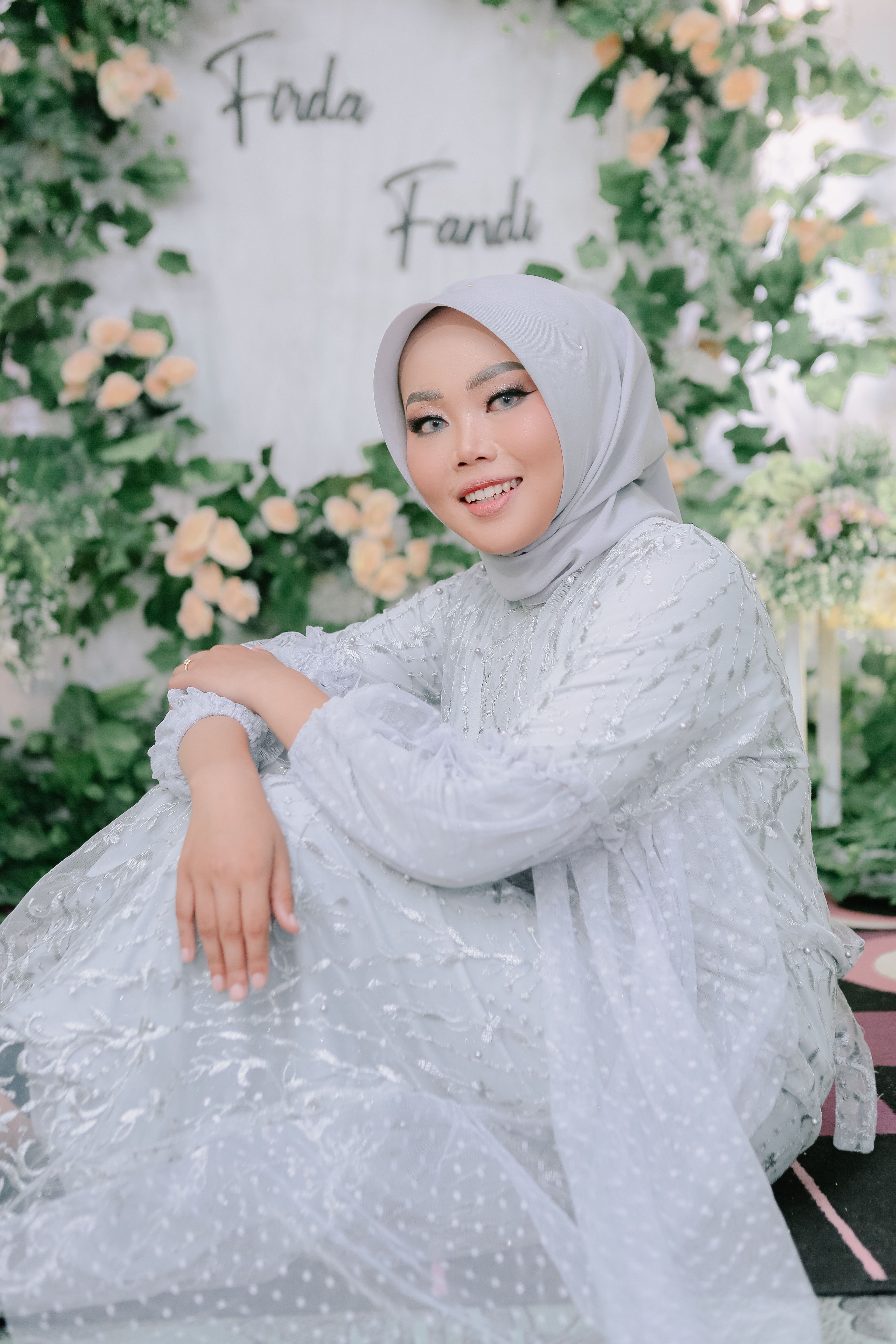 Smiling woman in a white hijab dress sitting surrounded by floral arrangement.