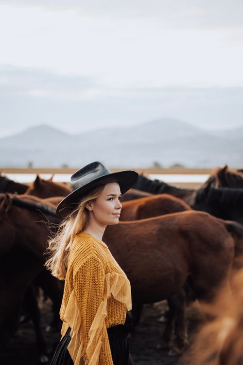 Free A woman in a stylish hat stands amidst a herd of horses against a stunning mountainous backdrop. Stock Photo