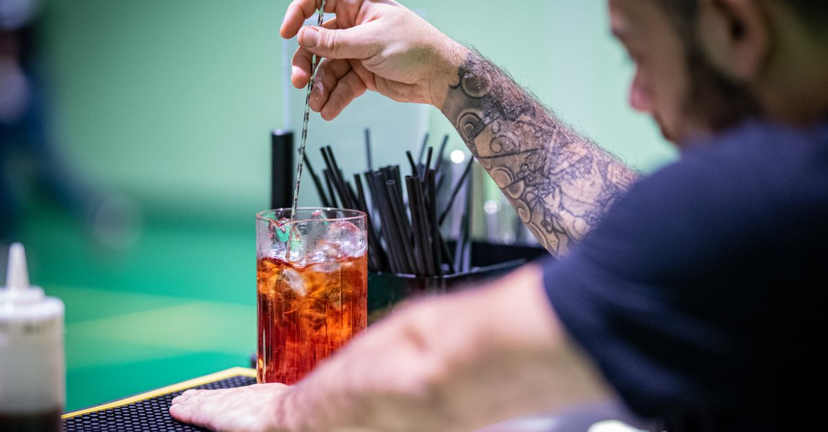 Tattooed bartender stirring a vibrant red cocktail with a mixing spoon at a bar.