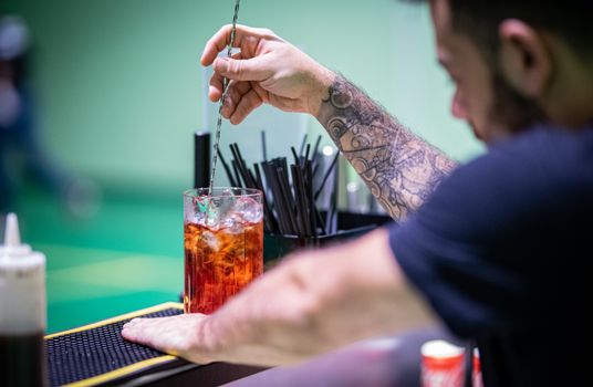 Tattooed bartender stirring a vibrant red cocktail with a mixing spoon at a bar.