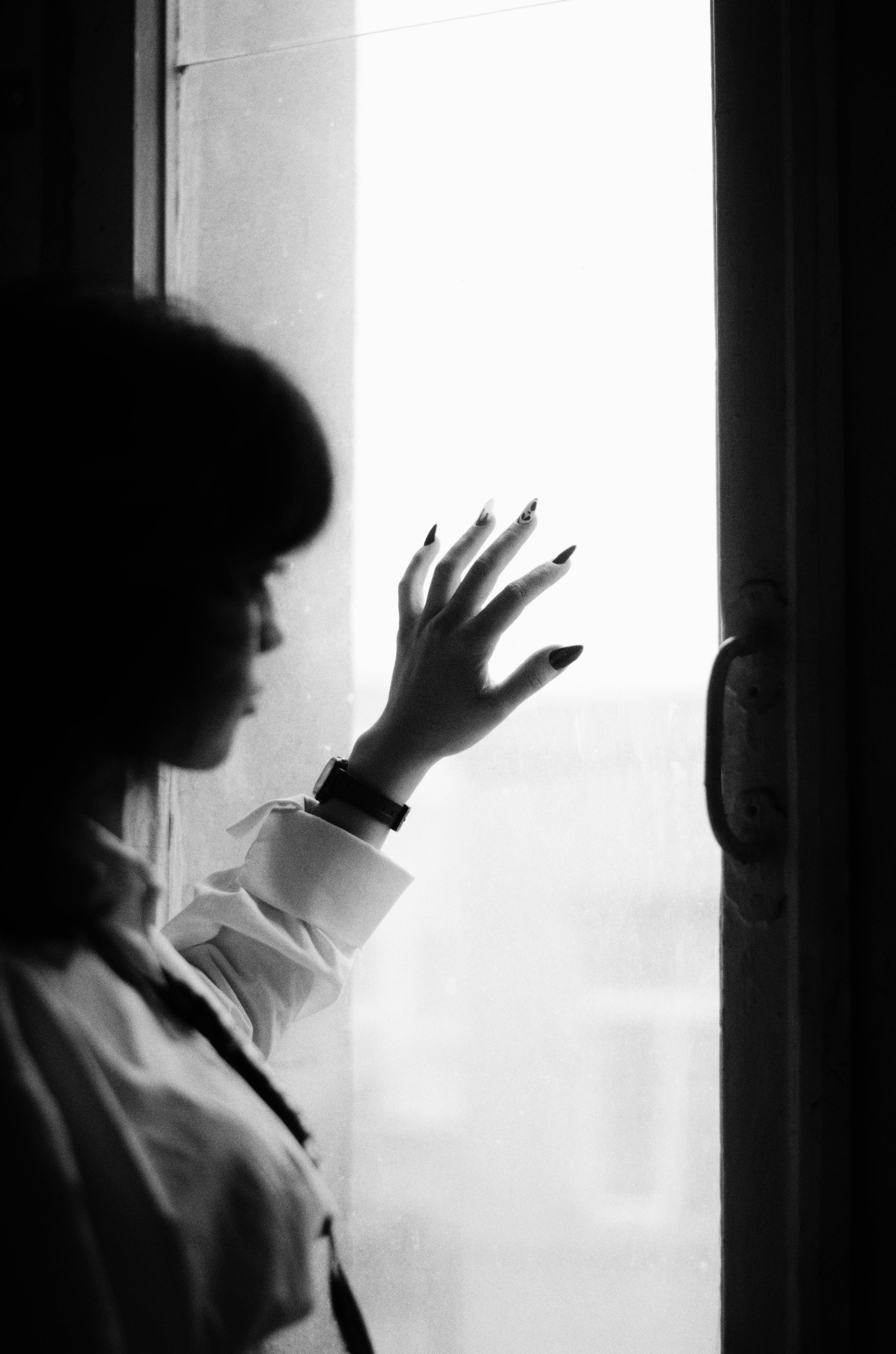 Black and white photo of a woman reaching towards a window, creating a silhouette effect.
