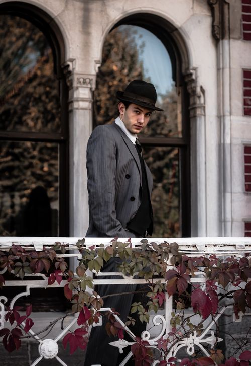 Free Elegant man in a suit and hat standing on a vine-covered balcony with classical architecture. Stock Photo