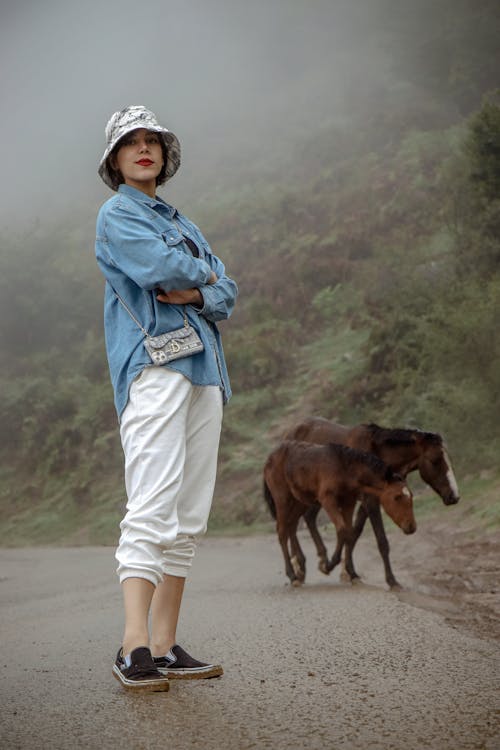 Woman Standing on a Road in Mountains with Two Horse Foals behind Her 