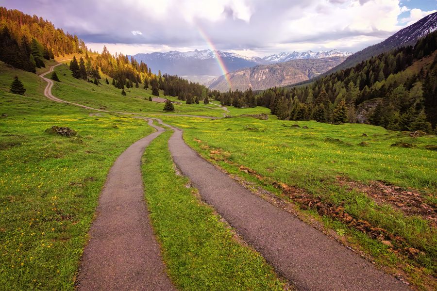 Scenic mountain path winding through lush greenery with a vibrant rainbow in the distance.