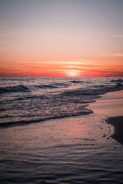 Free Calm ocean waves lap against the sandy shore as the sun sets on the horizon. Stock Photo