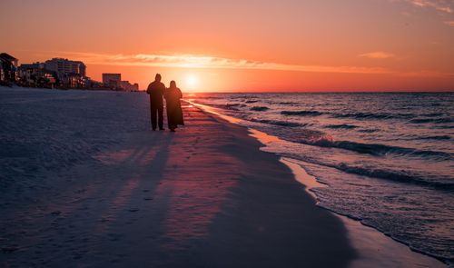 Free Two people walking along the beach at sunset, silhouetted against the colorful sky. Stock Photo