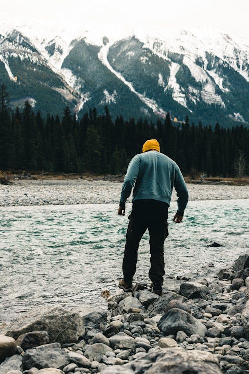 Man Standing on the Rocks near a Body of Water in a Valley with View of Trees and Mountains in the Background 