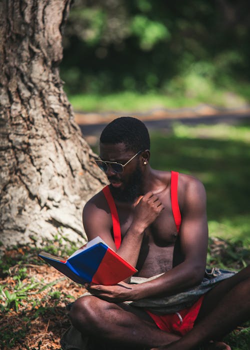 Topless Man Wearing Black Sunglasses Reading Book beside Brown Tree Trunk