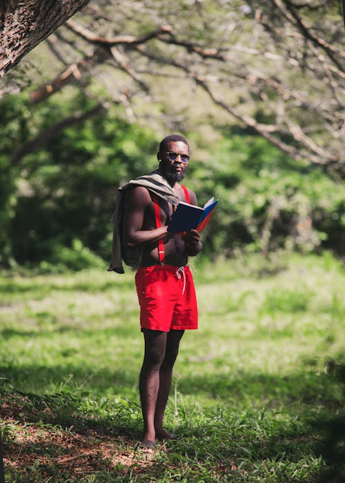 Muscular Man in Red Shorts Standing on Grass with Book in Hands