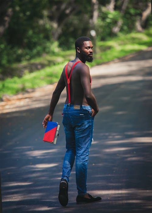 Shirtless Man in Blue Denim Jeans Walking on Asphalt Road Carrying a Book 