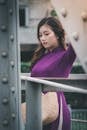 一位身穿紫色连衣裙的年轻女子头戴编织帽，在城市金属桥上优雅地摆着姿势